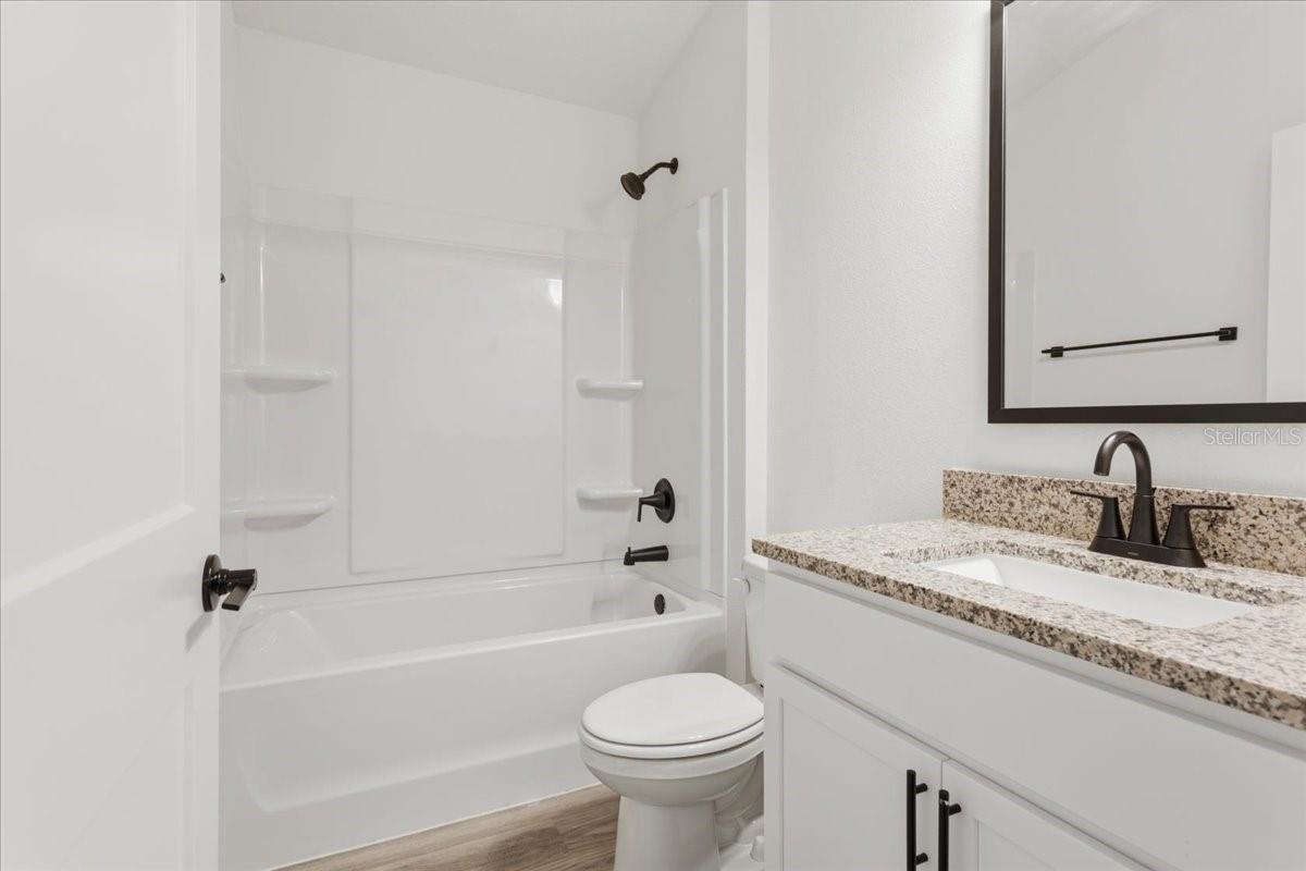 A modern bathroom featuring a white vanity with granite countertop, matte black faucet, mirror with black frame, toilet, and a bathtub-shower combo with white walls.