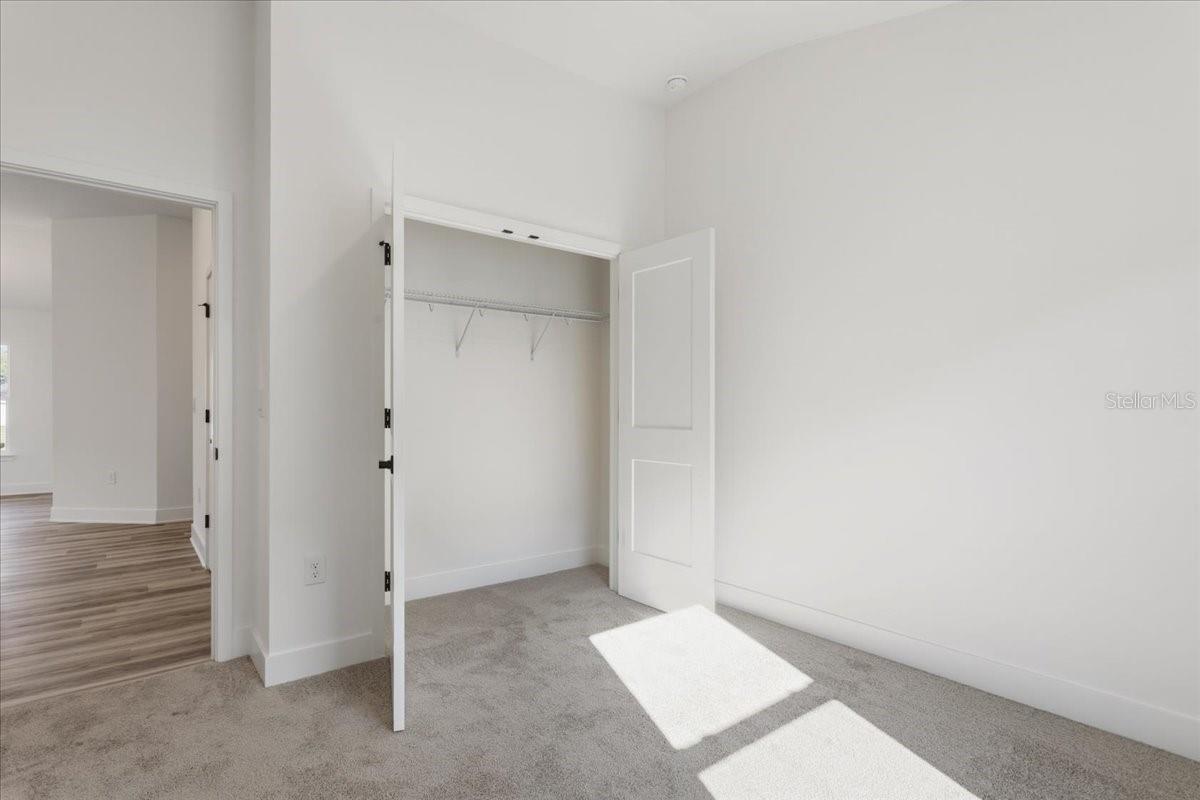 An empty bedroom with gray carpet, white walls, and an open closet door revealing a wire shelf.