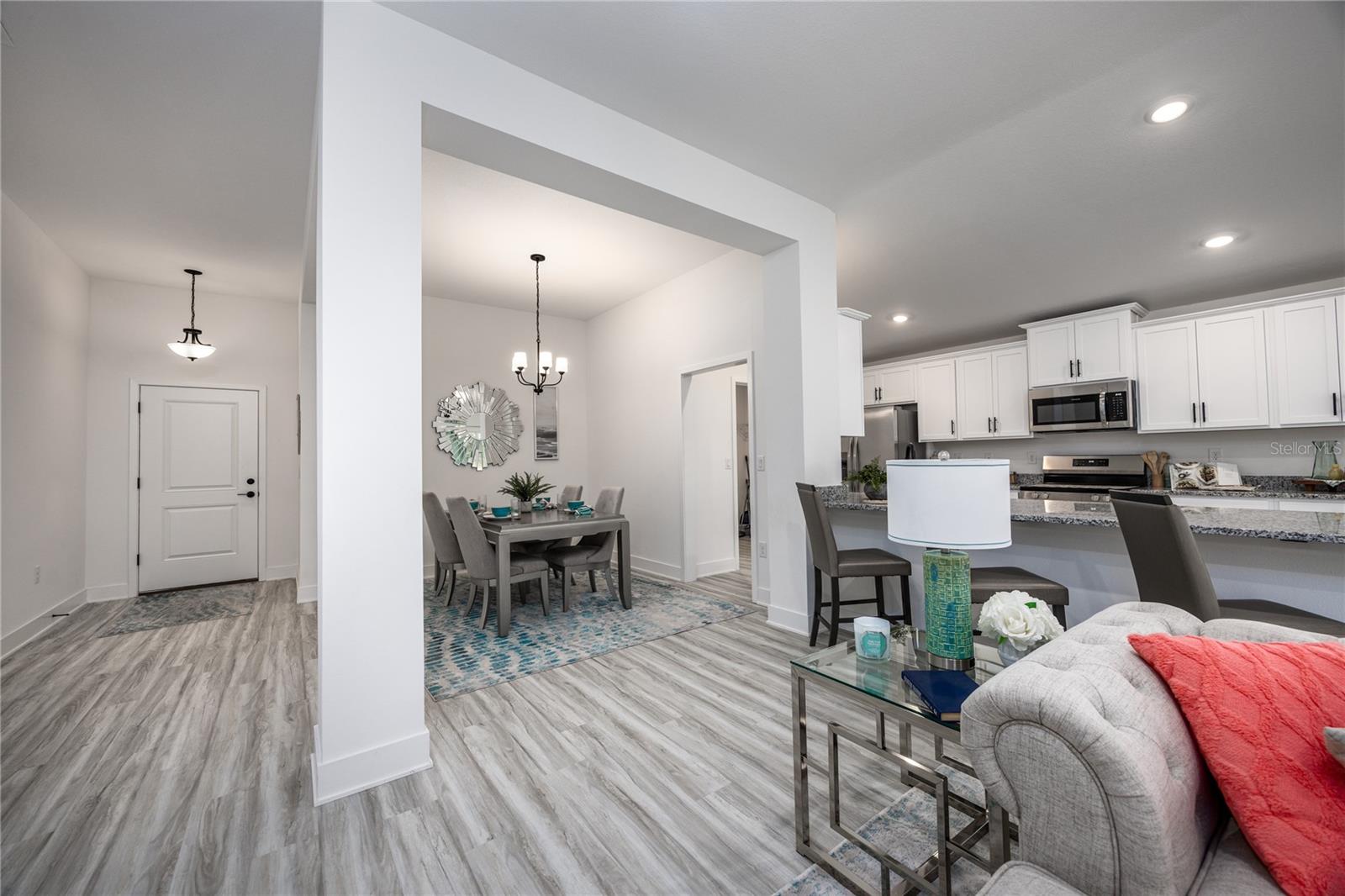 A wide angle shot showing the open concept living, dining, and kitchen area with white cabinets and grey wood-look flooring.