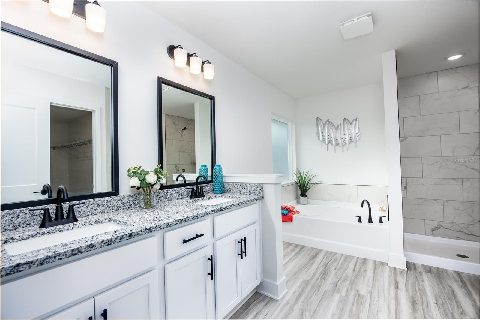Modern master bathroom featuring a white double vanity with granite countertop, black fixtures, and a soaking tub next to a walk-in shower.