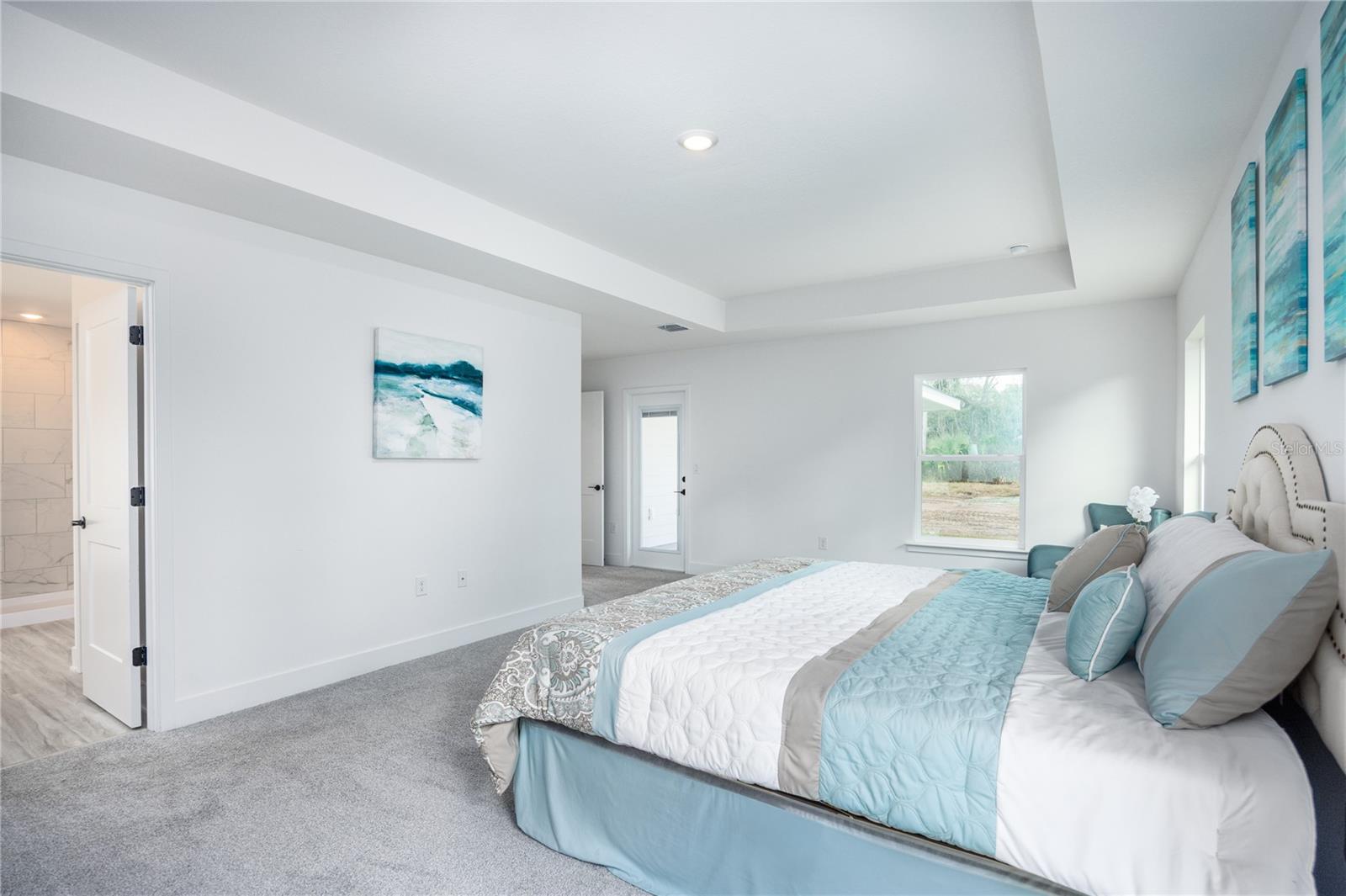 A modern master bedroom with a king-sized bed featuring blue and grey bedding, a tray ceiling, and a doorway leading to a tiled bathroom.