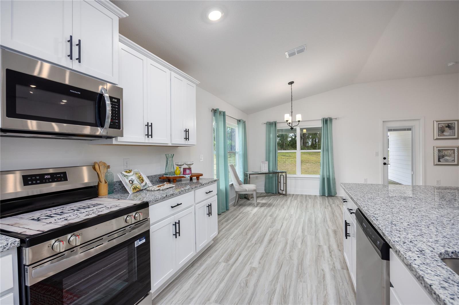 Bright, modern open-concept kitchen featuring white cabinets, granite countertops, stainless steel appliances, and a connected dining area with teal curtains.