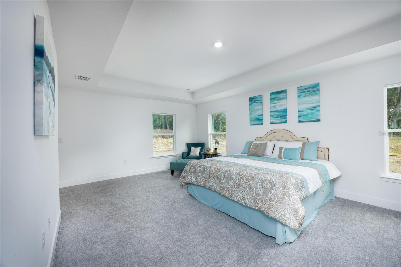 Spacious master bedroom with light gray carpet, a bed with light blue and grey bedding, a blue armchair in the corner, and three abstract blue paintings above the headboard.