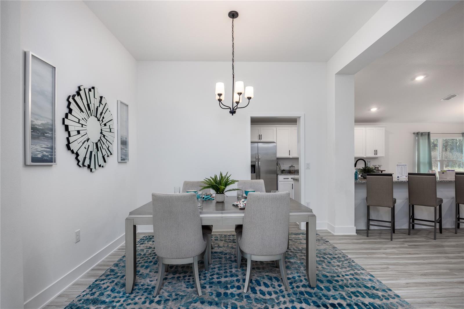 A modern dining room with a rectangular table, four grey chairs, a blue patterned rug, and a modern chandelier, leading into a kitchen.