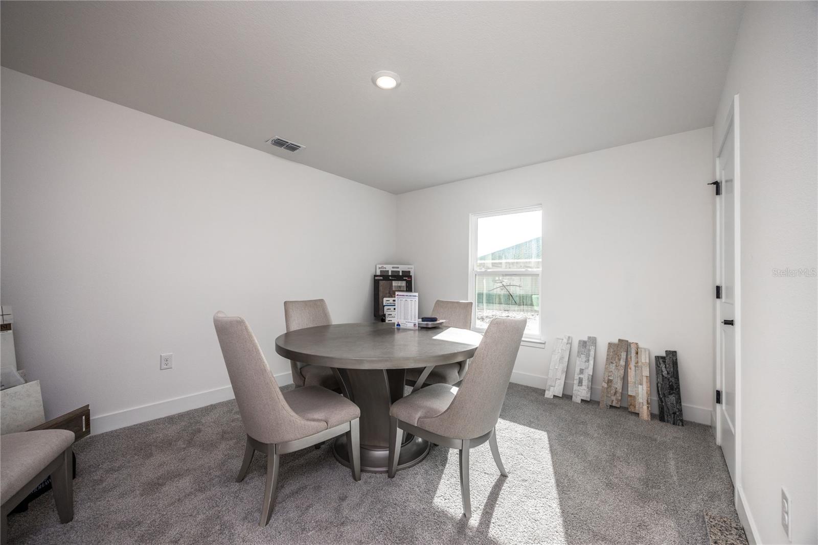 A small dining area with a round table, four upholstered chairs, grey carpet, and plain white walls in a new construction setting.