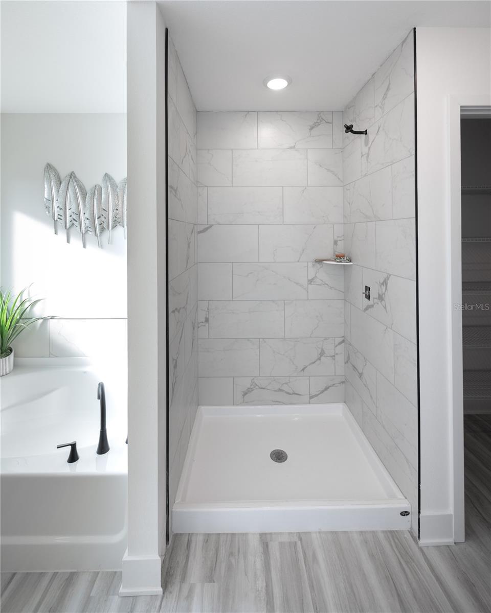 A modern shower with white marble-look tiles, a black showerhead, and a adjacent bathtub with decorative wall art in a brightly lit bathroom