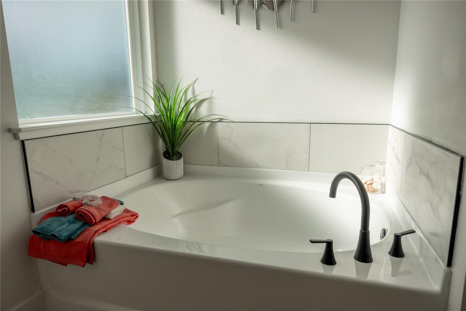 Corner soaking tub in a bathroom featuring white marble-look tiles, a frosted window, a small decorative plant, and colorful towels.