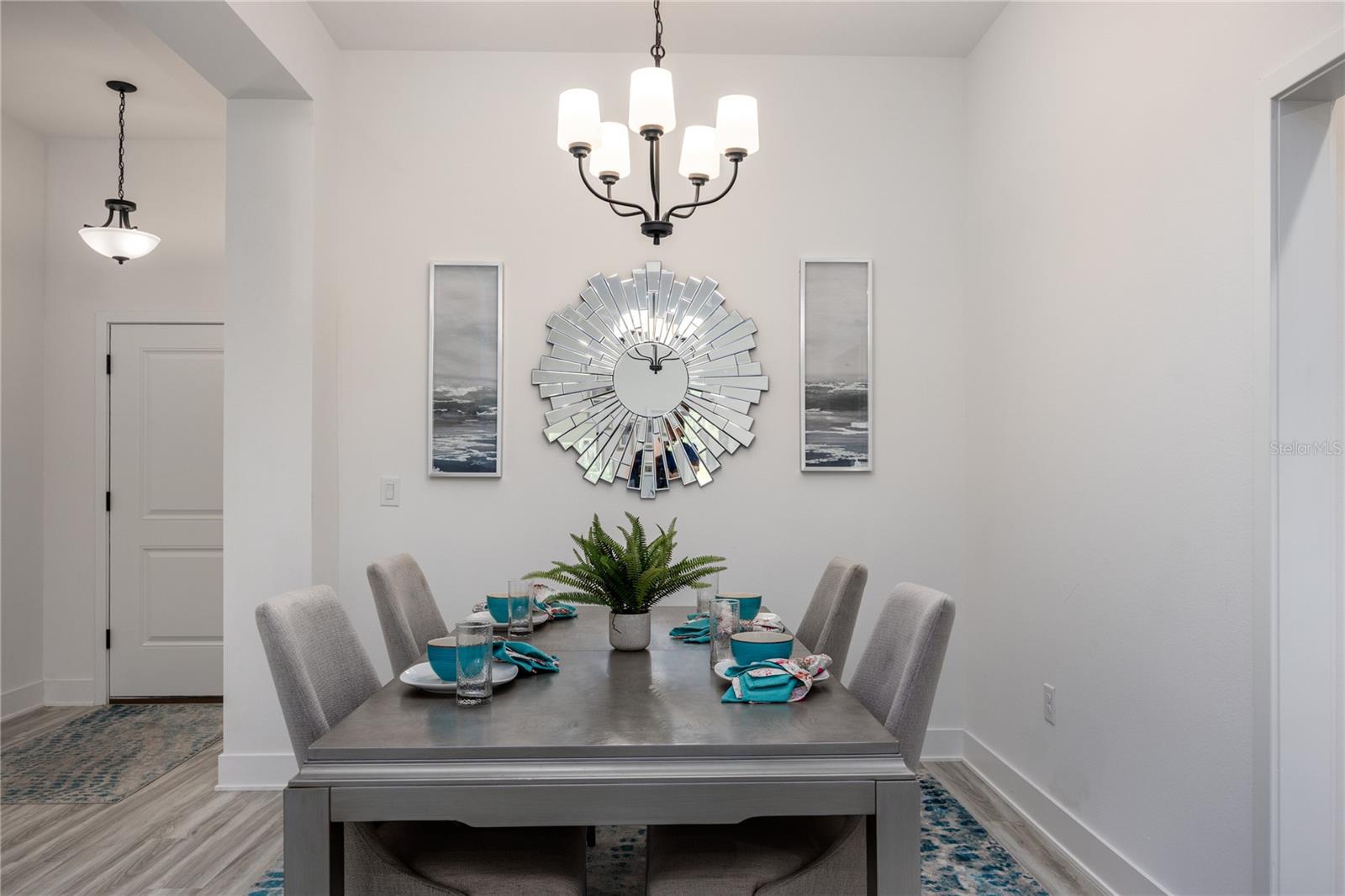 A dining room featuring a grey table, upholstered chairs, a decorative wall mirror, and modern lighting.