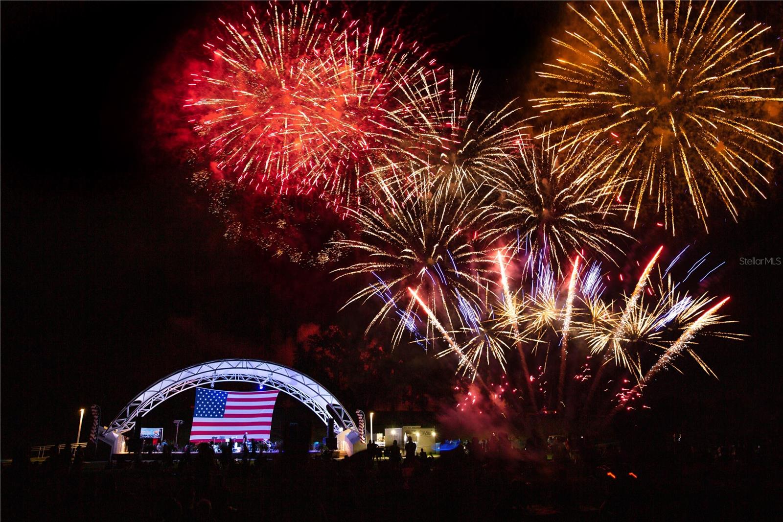 A festive night scene with colorful fireworks bursting in the sky above an outdoor stage featuring an American flag backdrop, representing community events.