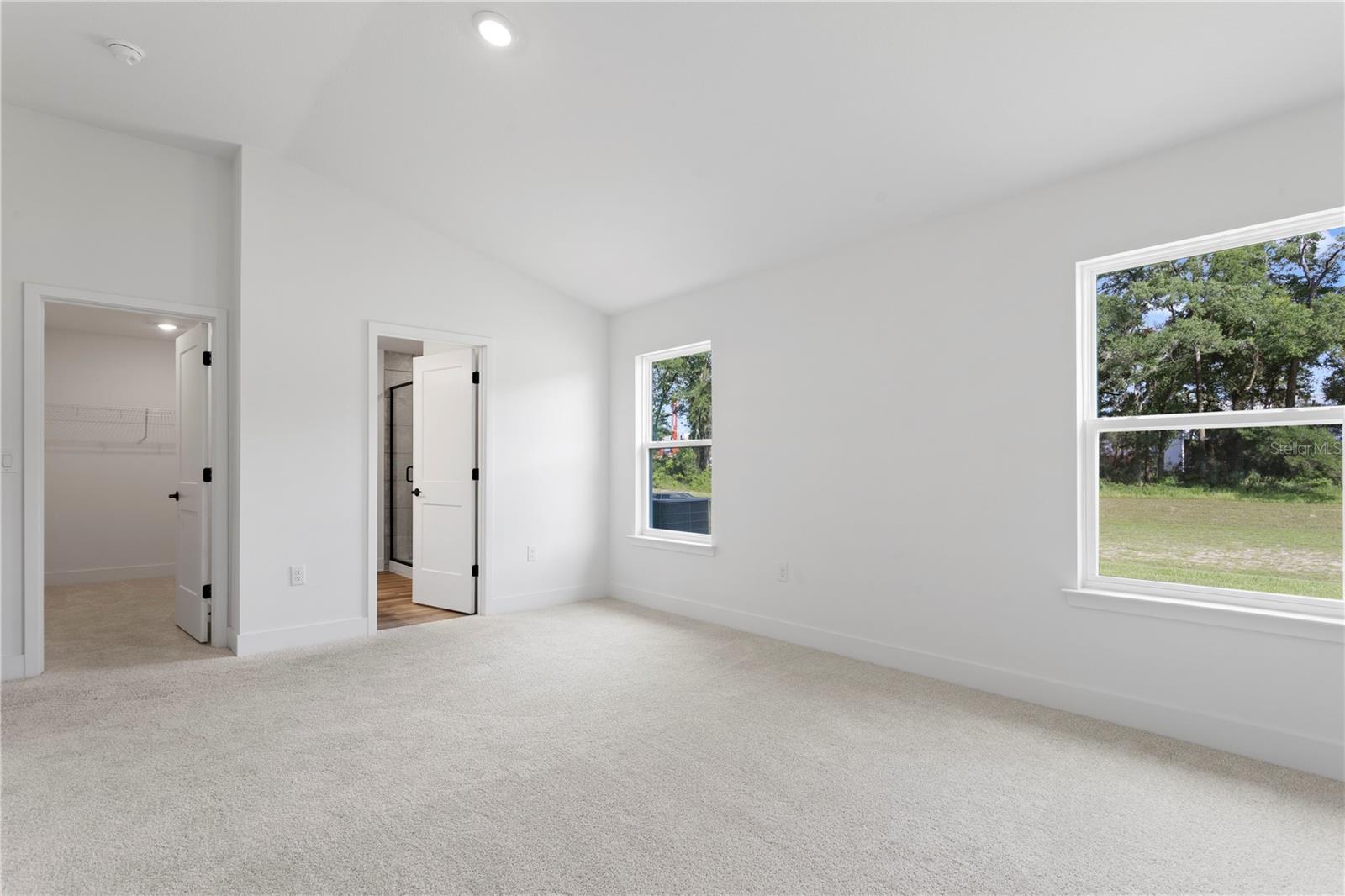 A vacant bedroom with neutral carpet and white walls featuring two windows, a doorway to an ensuite bathroom, and a closet door