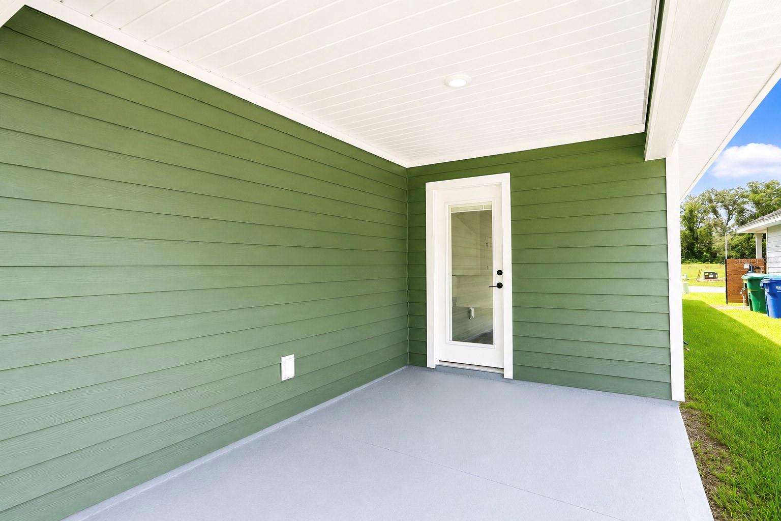 A covered concrete patio with olive green siding and a glass door leading into the home.