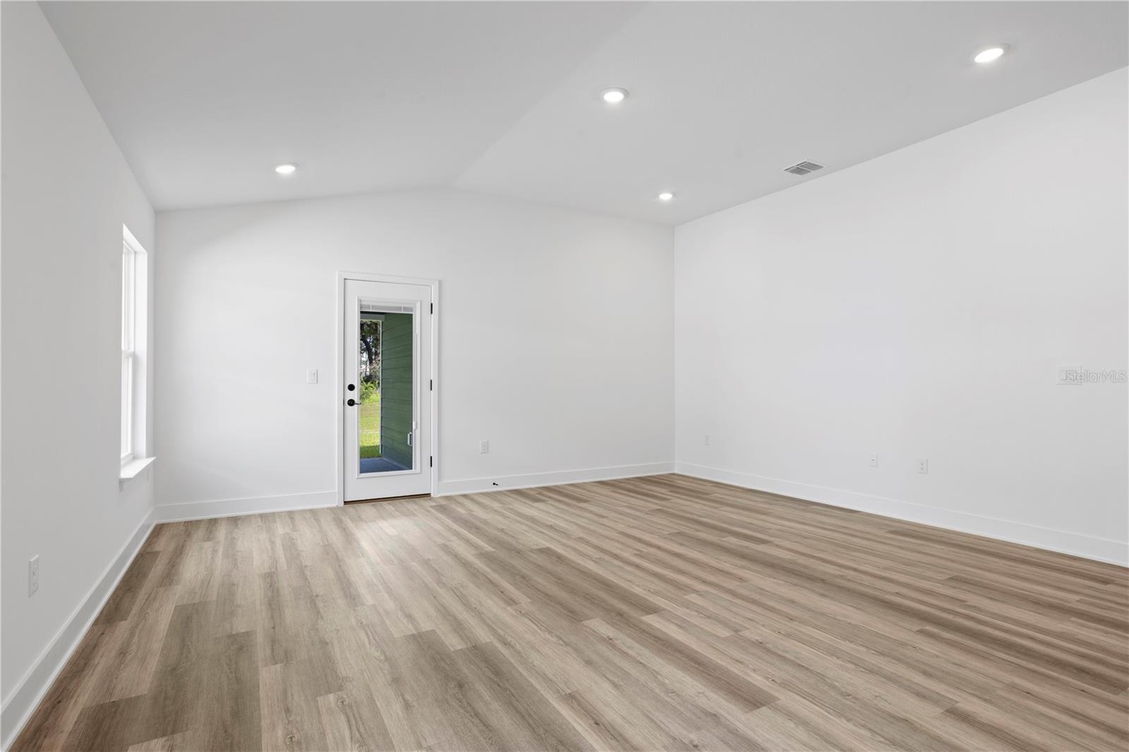 A wide angle shot of a vacant living room with white walls, light wood-look luxury vinyl plank flooring, recessed lighting, and a glass-paneled exterior door.