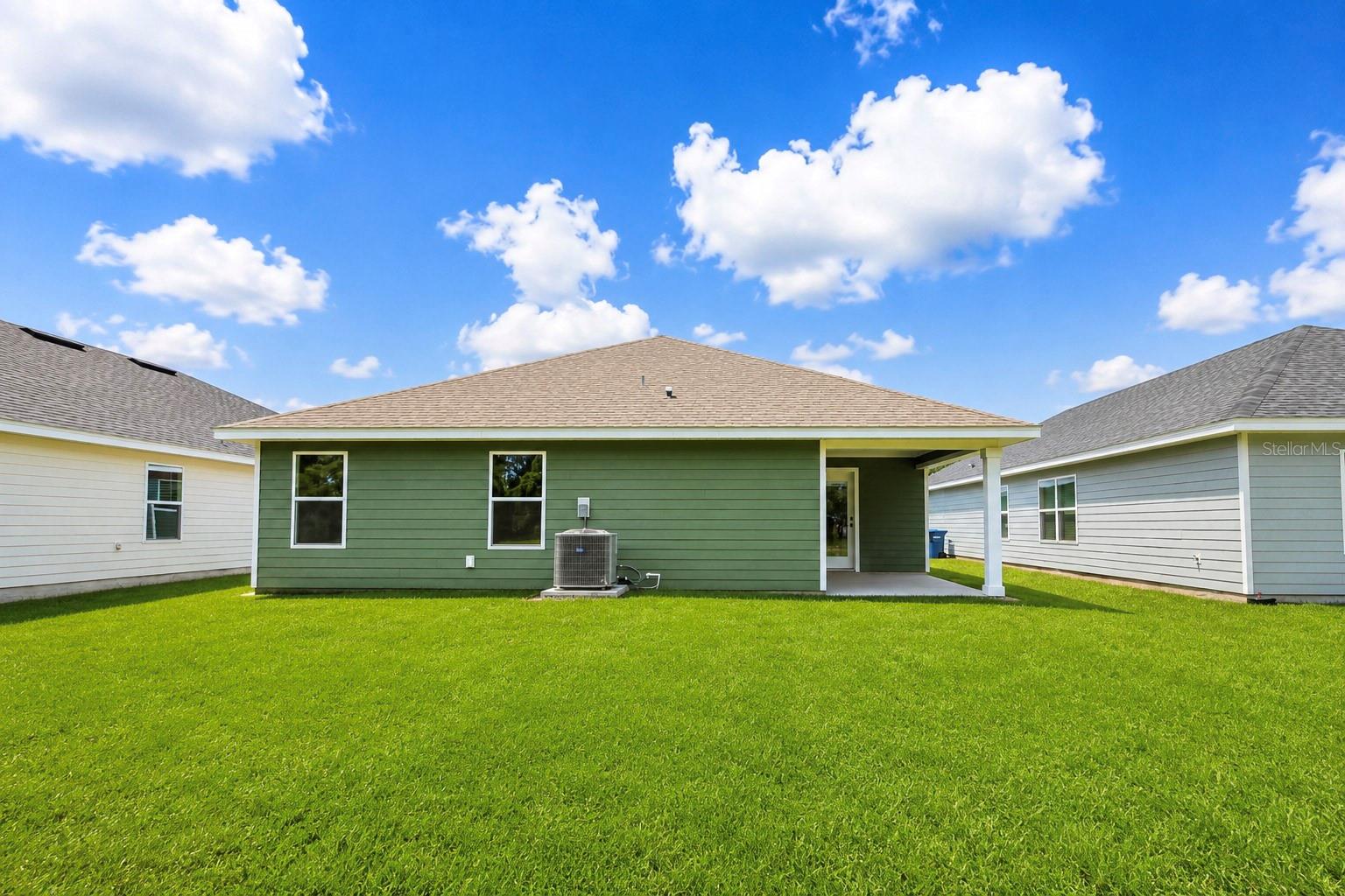 The back exterior of a green single-story house with a lawn, a small concrete patio, and an HVAC unit under a bright blue sky.