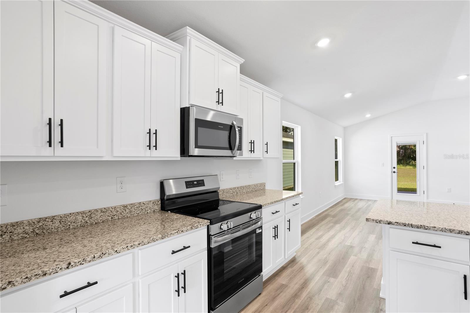 Modern kitchen with white cabinetry, granite countertops, and stainless steel appliances, featuring wood-look flooring and an open layout with a view of a doorway.