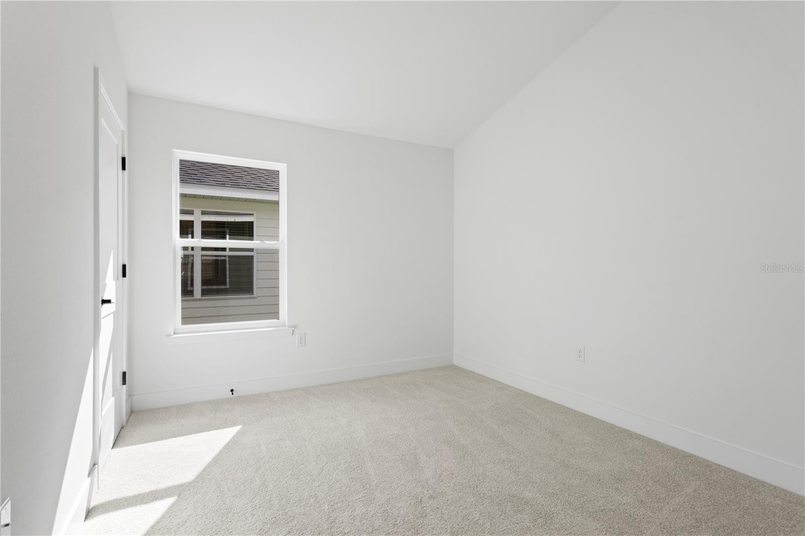 An empty bedroom with white walls, carpeted flooring, and a window.