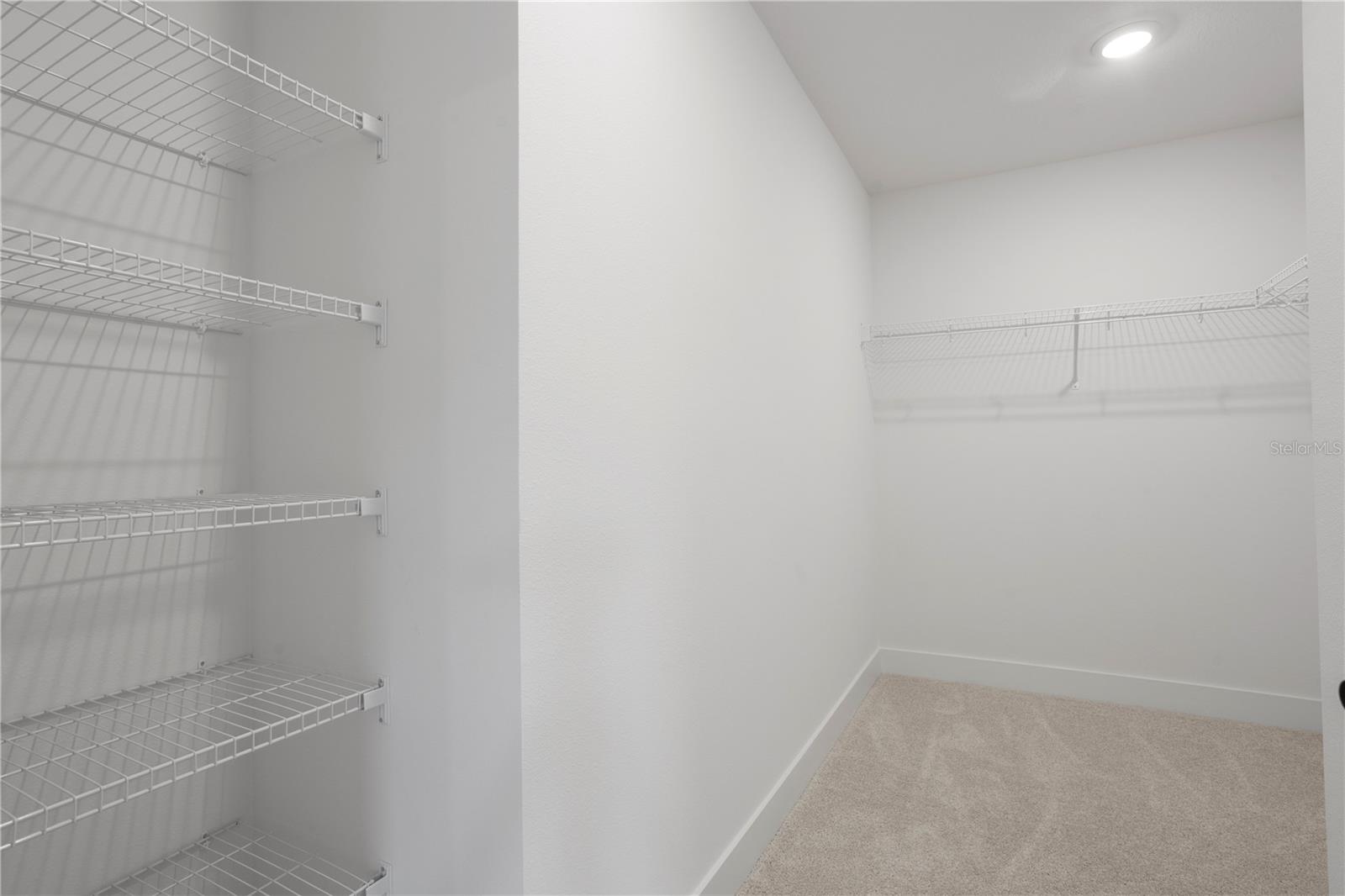A walk-in closet featuring white wire shelving units and tan carpeting.