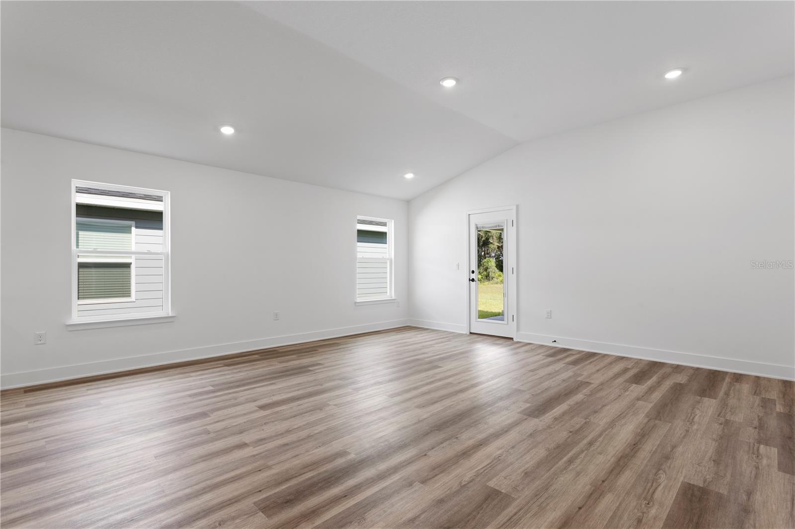 Bright living room with white walls, LVP wood-style flooring, vaulted ceiling, recessed lighting, and a glass door leading outside.