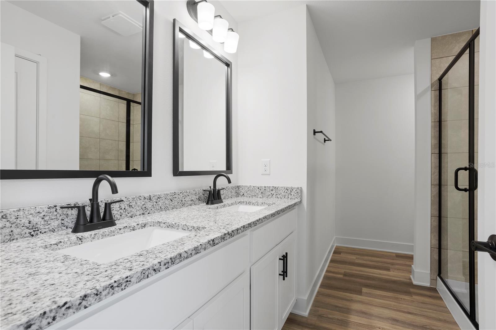 A modern bathroom vanity with dual sinks, speckled granite countertop, black fixtures, and a walk-in shower.