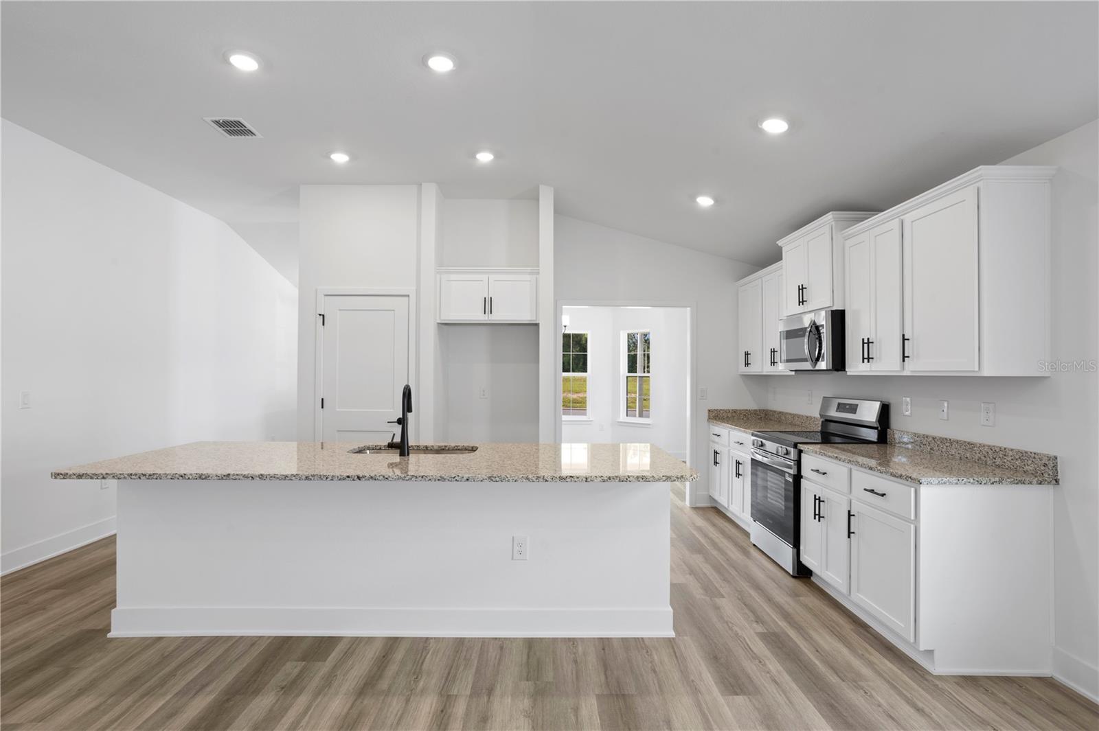 A modern kitchen featuring a large white center island with a sink, white cabinetry, granite countertops, and light wood-tone flooring.