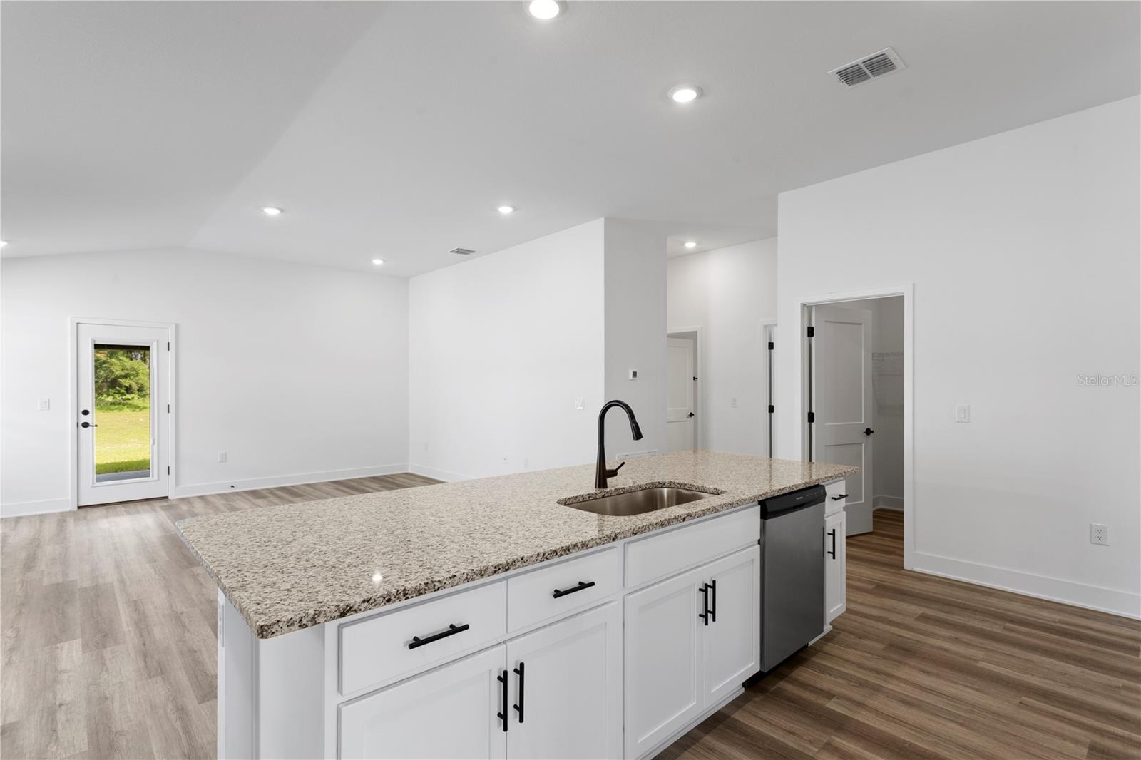 A white kitchen island with granite countertops and a stainless steel sink sits in an open-concept living space with white walls and light wood plank flooring.