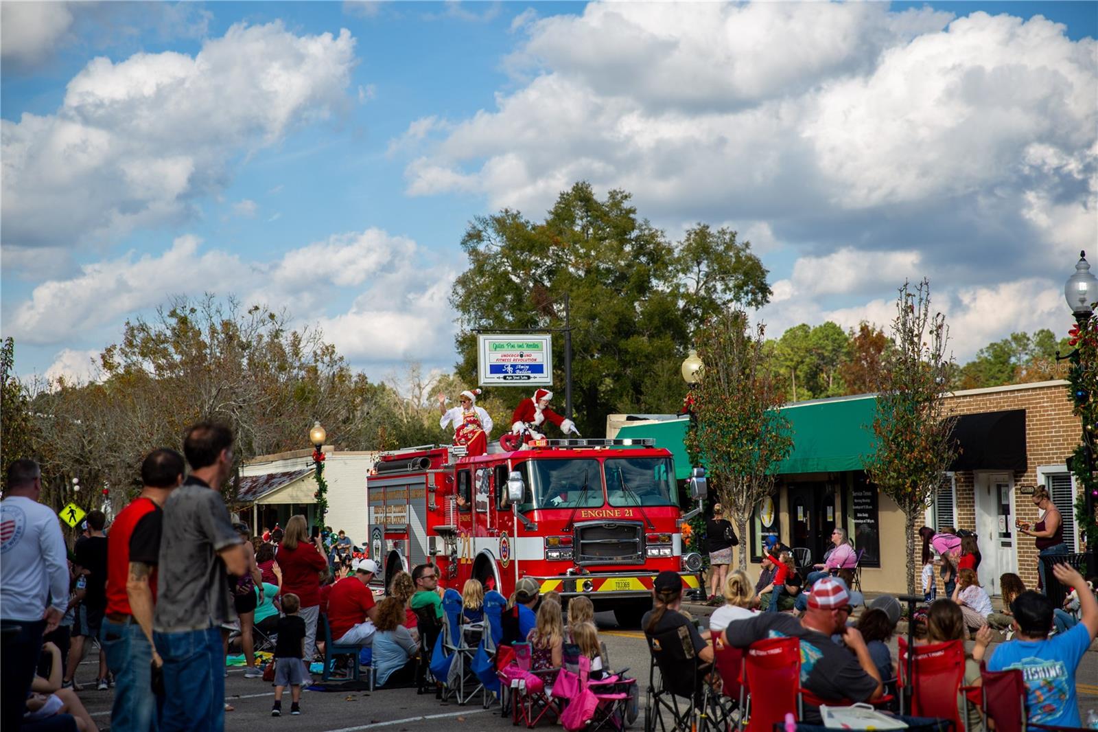Santa Claus waving from a fire truck in a local parade with a crowd of spectators in folding chairs.