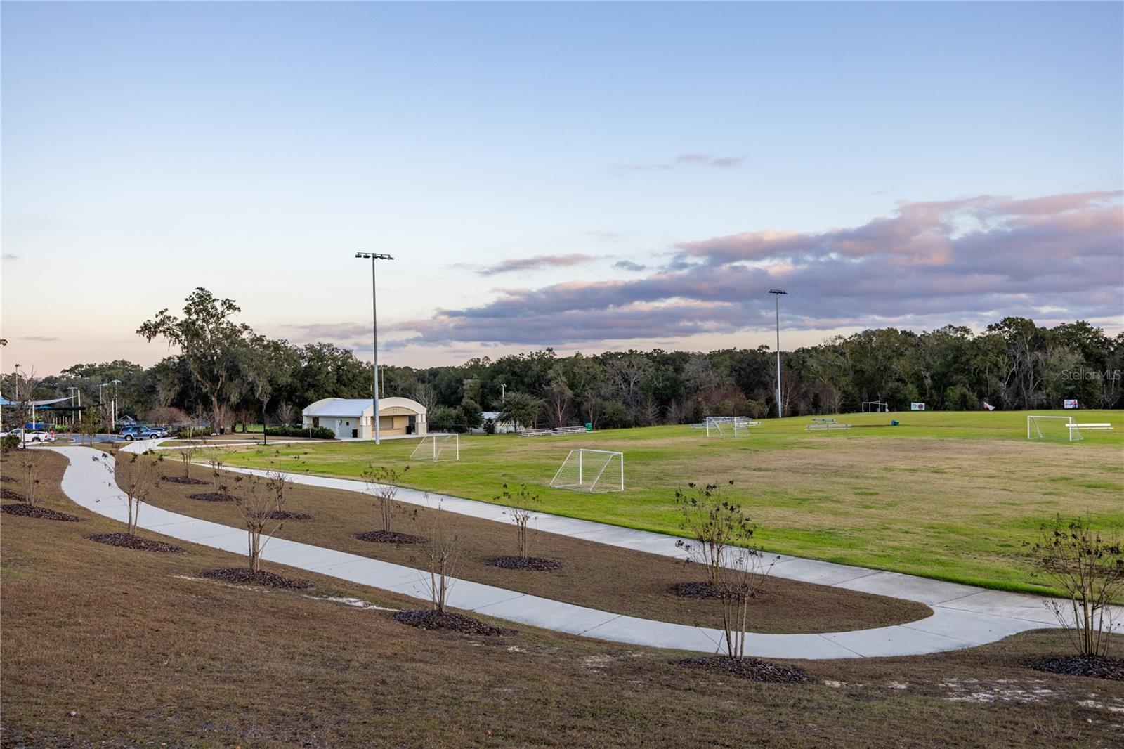 A community soccer field with walking paths, stadium lighting, and a pavilion under a clear sky.