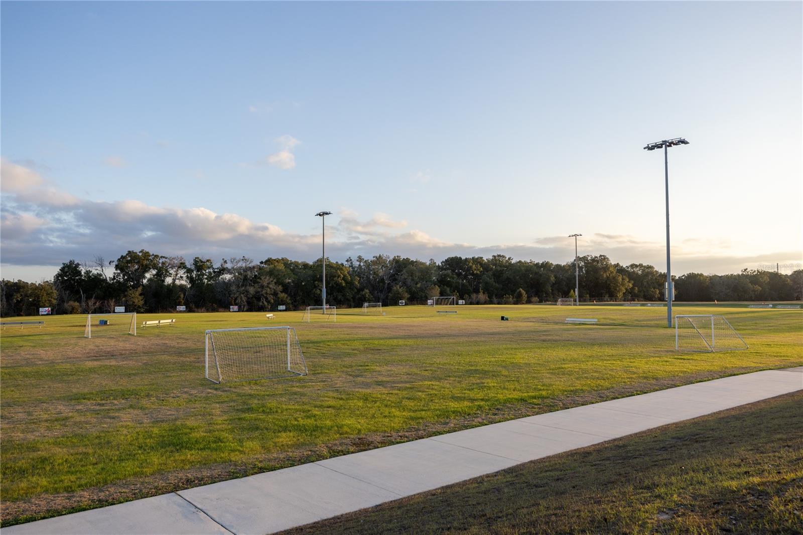 A wide view of an outdoor community soccer field with multiple goals and light poles at sunset.