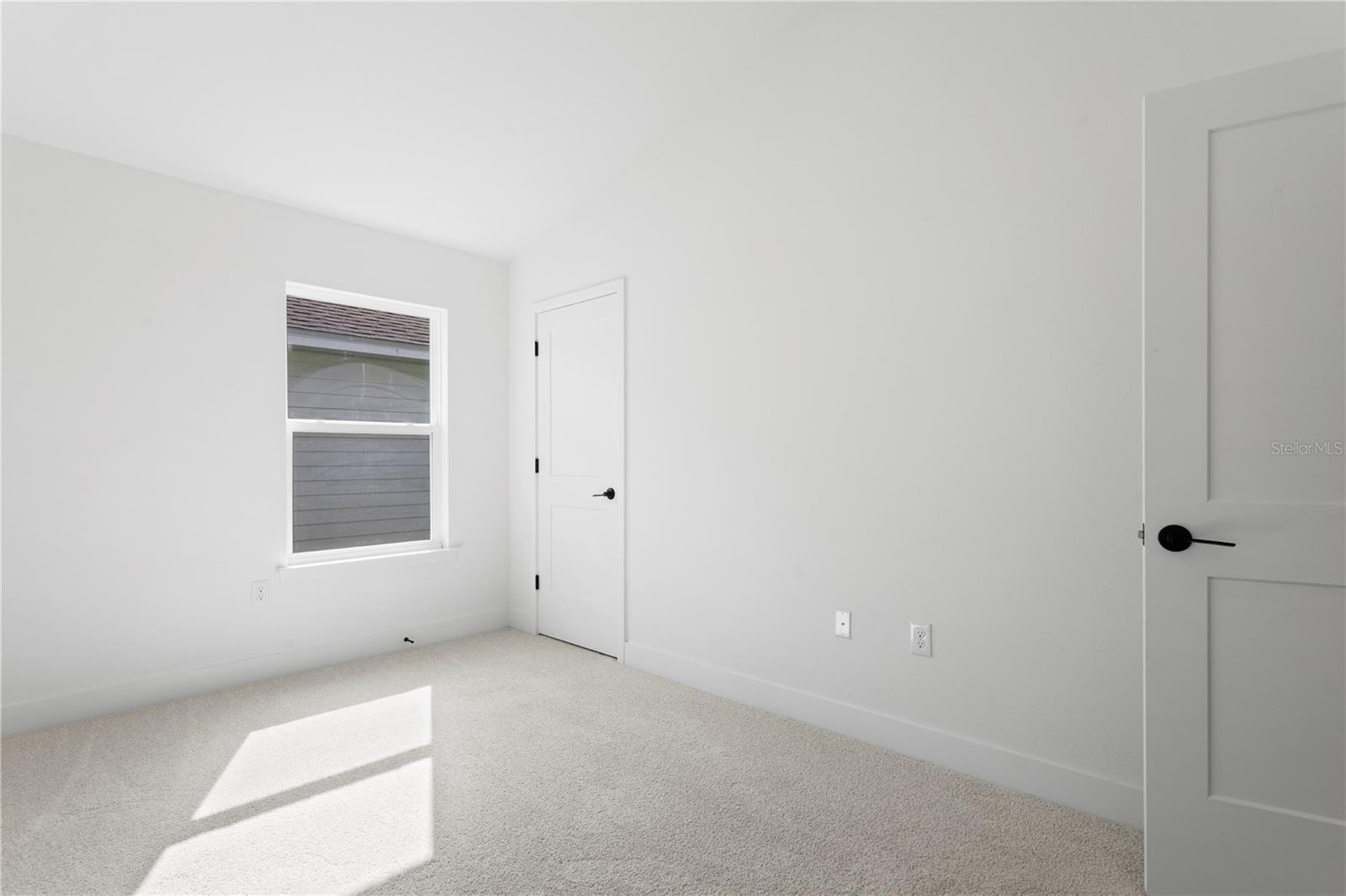 A vacant bedroom with white walls, beige carpet, and a single window.