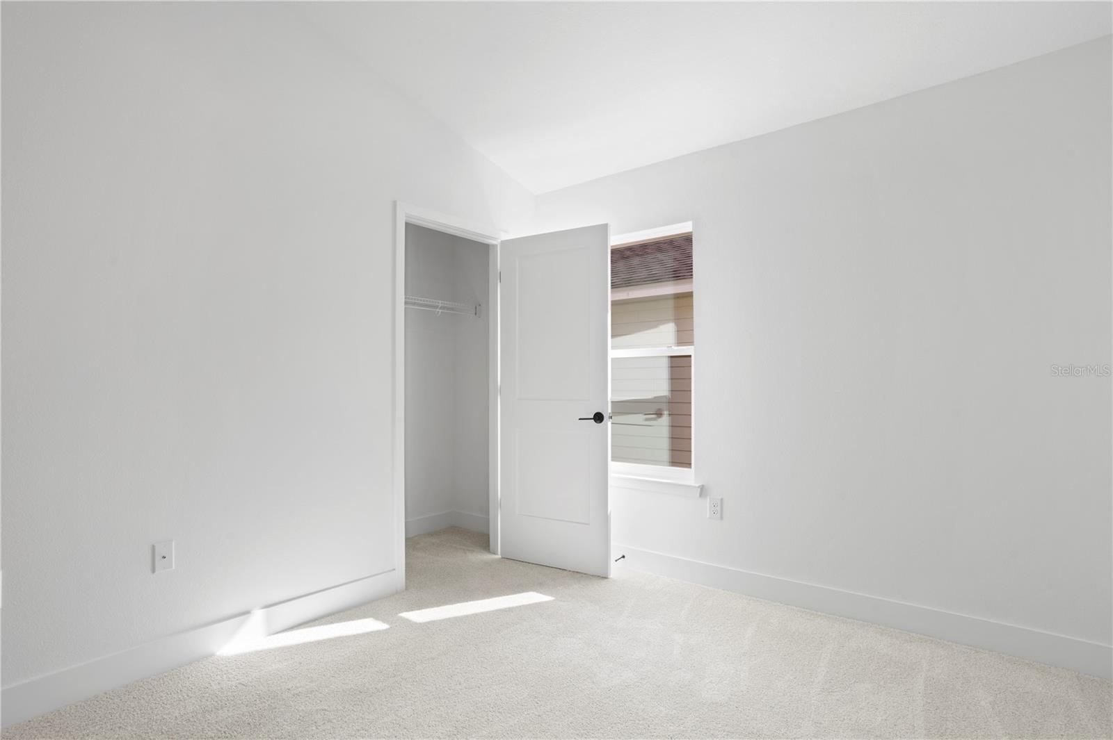 A sparse, bright bedroom with white walls, beige carpet, and an open closet door.