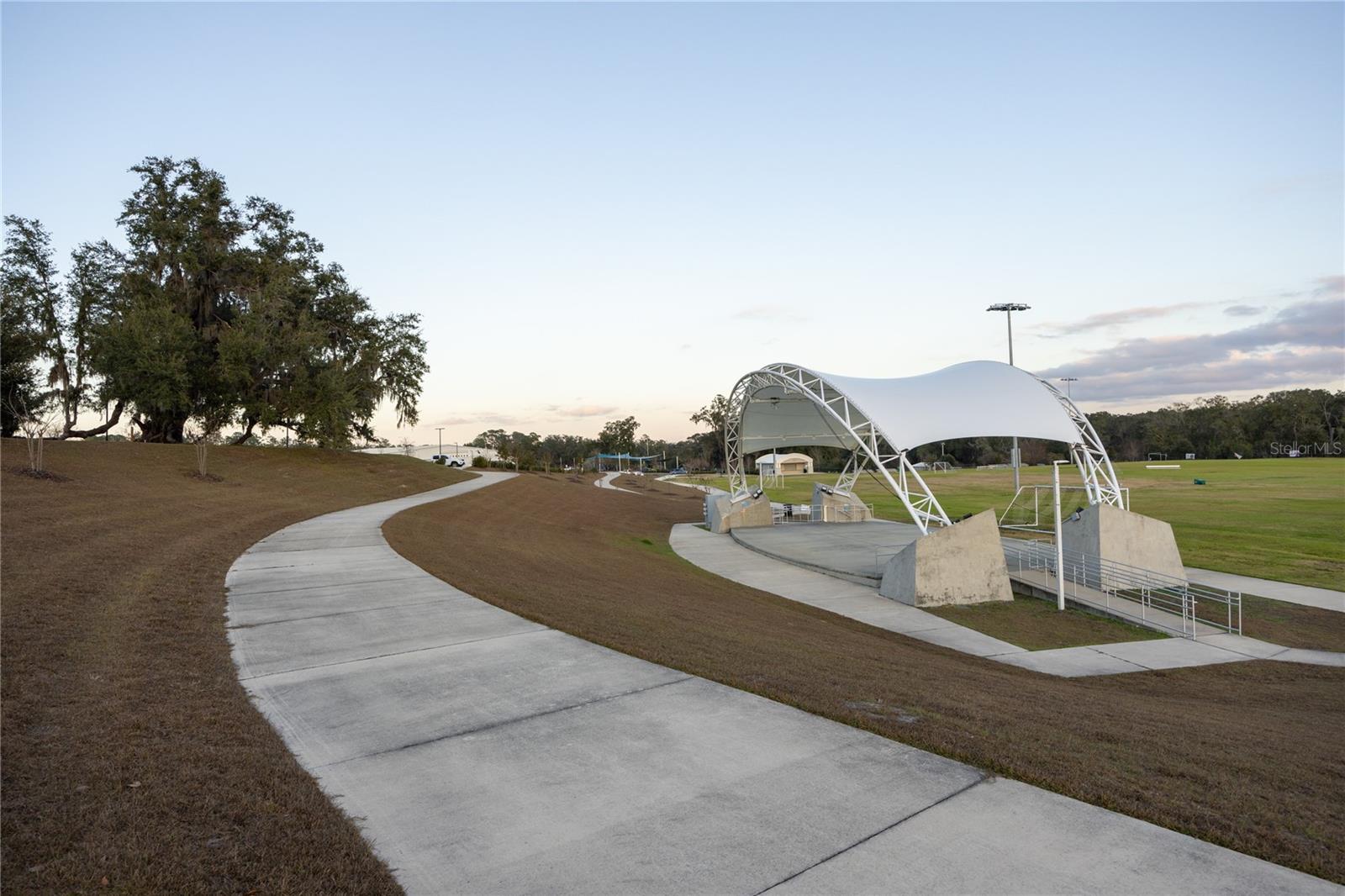A paved walking path leads toward a modern, white tensile roof amphitheater in a community park setting during late afternoon.