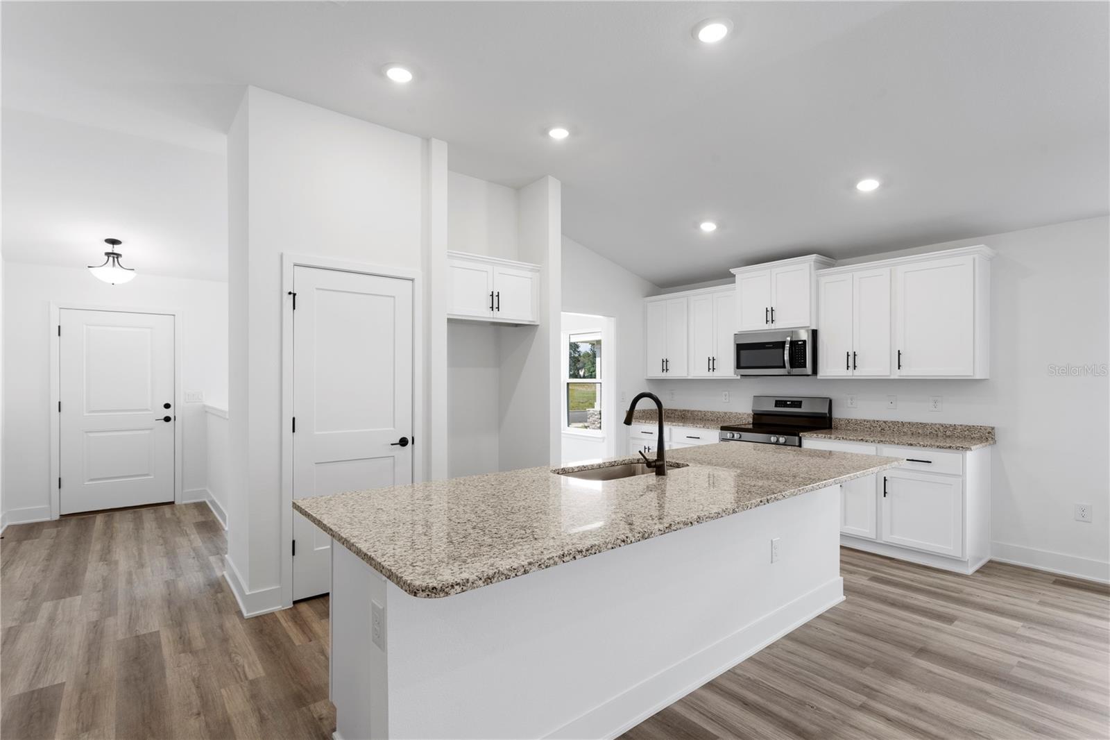 A modern, bright, white-cabinet kitchen with a granite island, recessed lighting, and light wood-look flooring.