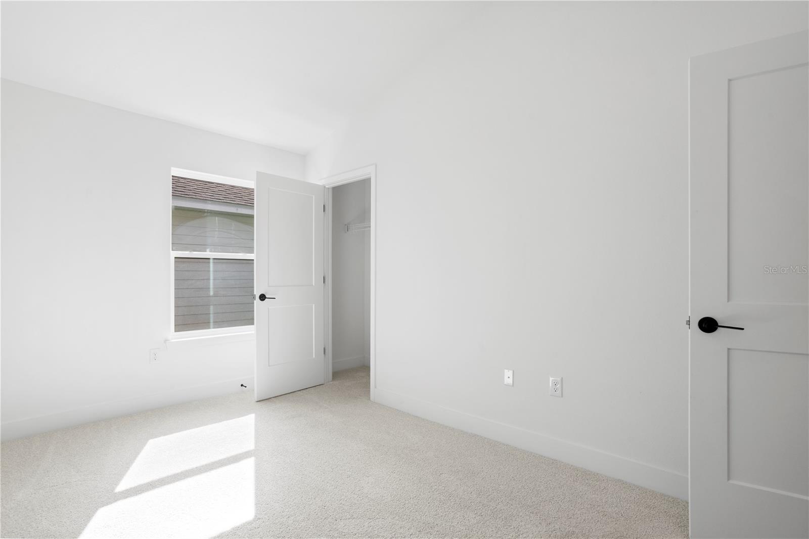 A clean, empty bedroom with white walls, carpeted floors, a window, and an open closet door.