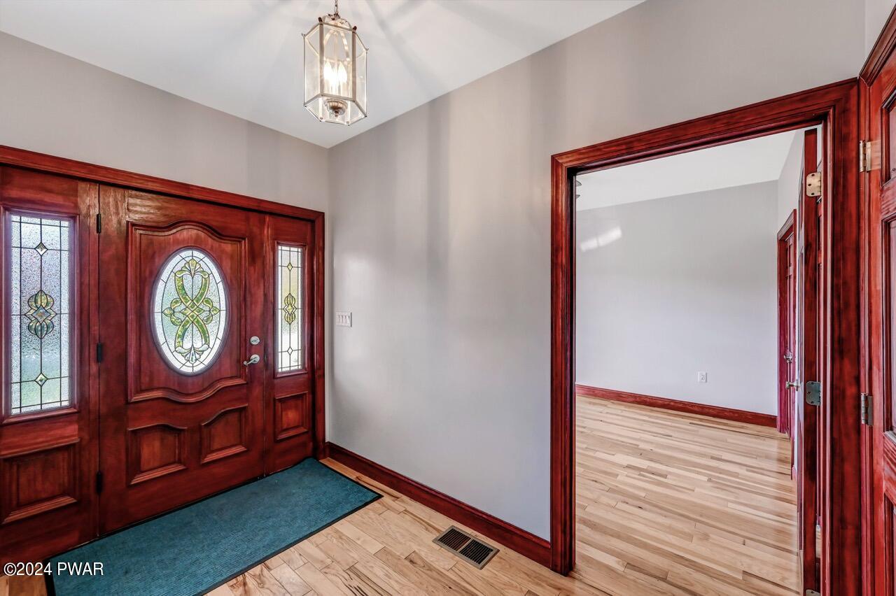 Entry foyer with stained wood front door featuring decorative glass panels, hardwood floors, gray walls, a hanging light fixture, and a doorway to an adjacent room.