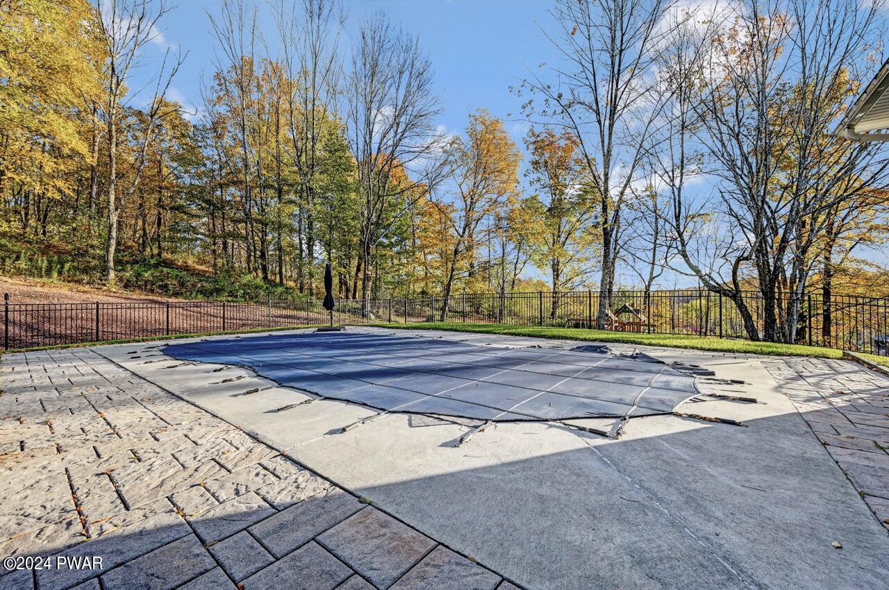 Outdoor stone patio overlooking a covered swimming pool, surrounded by a black metal fence and autumn trees.