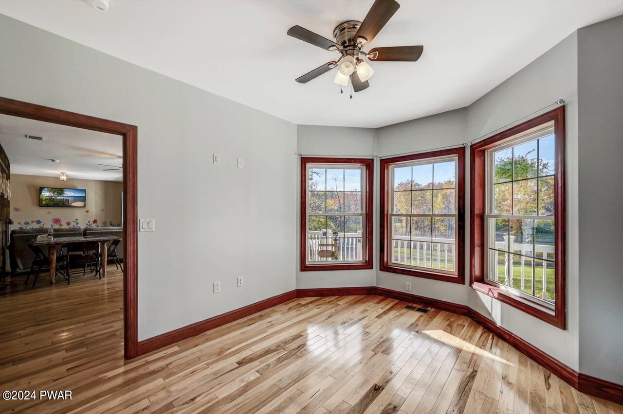 Empty sunroom with three bay windows, hardwood floors, gray walls, wood trim, and a ceiling fan; doorway opens to a dining area.