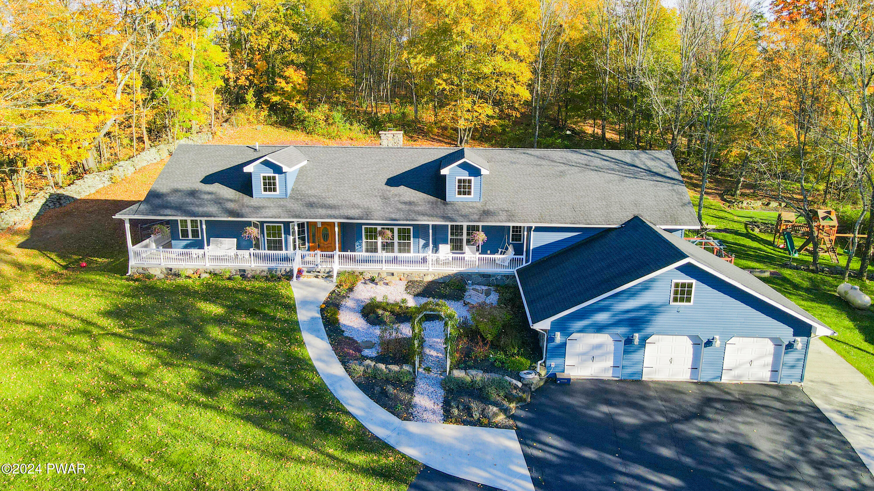 Aerial view of a blue house with a front porch, detached three-car garage, curved walkway, and autumn trees surrounding the property.