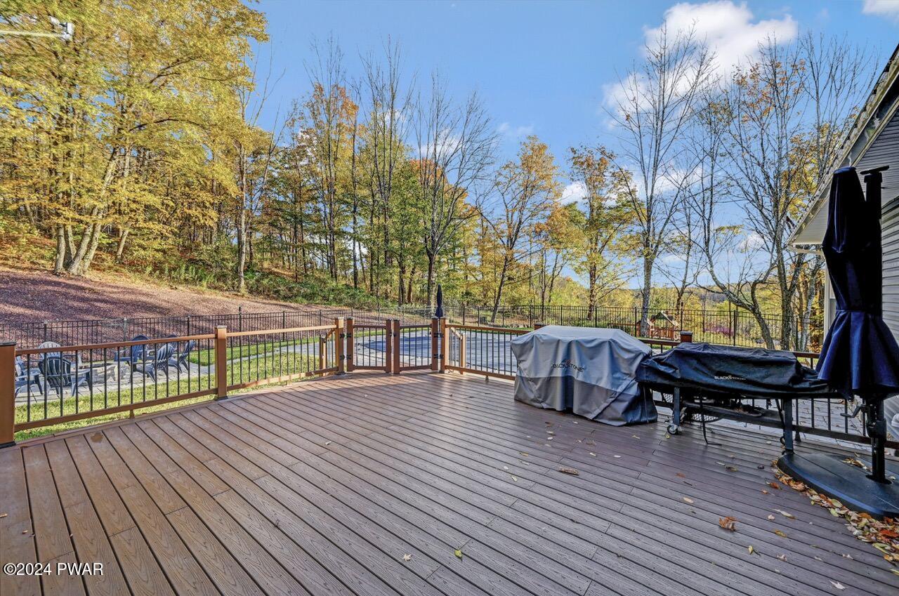 Wooden backyard deck with railing, covered grill and patio furniture, overlooking a fenced yard with a pool and autumn trees.