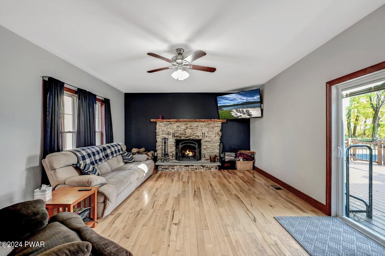 Living room with light wood floors, a stone fireplace on a dark accent wall, a sofa along the left wall, a ceiling fan, and a sliding glass door opening to a deck and pool area.