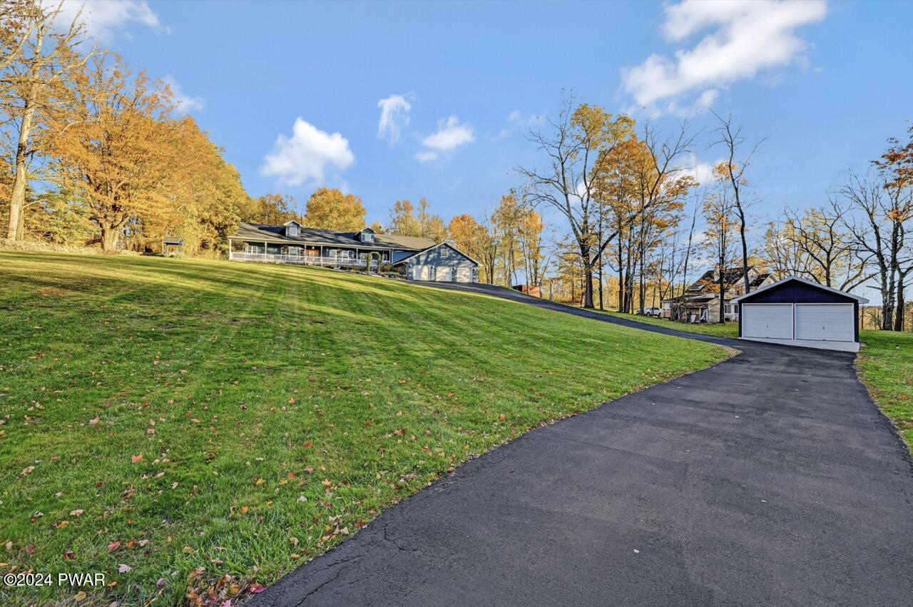 Wide exterior view of a home set on a sloped grassy yard with a long paved driveway leading to a detached two-car garage, surrounded by trees in autumn.