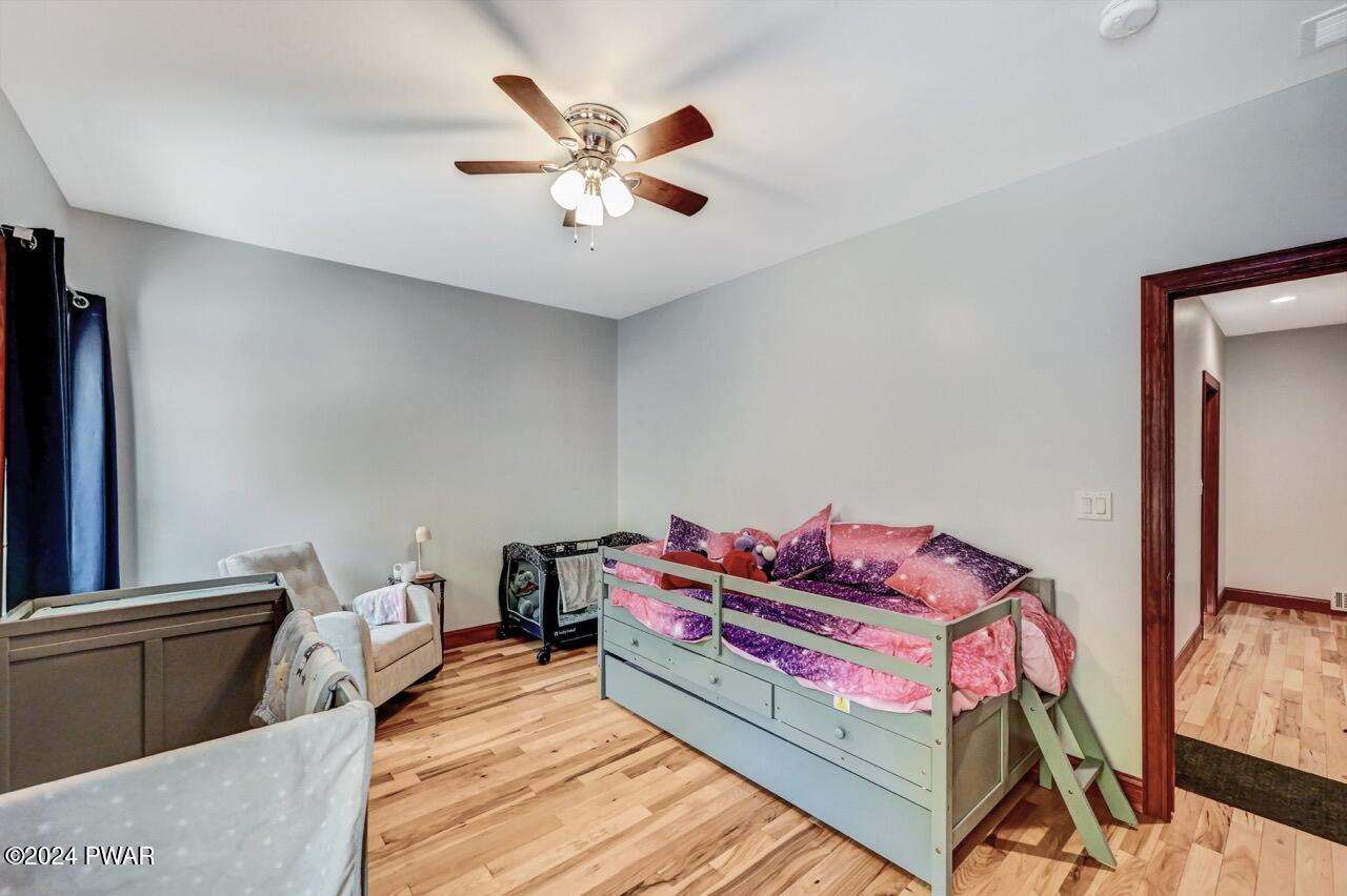A bedroom with light gray walls, hardwood floors, a ceiling fan, a daybed with colorful bedding, an armchair, and a hallway doorway.