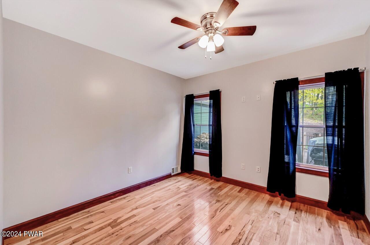 Empty bedroom with hardwood floors, light gray walls, two windows with dark curtains, and a ceiling fan with lights.