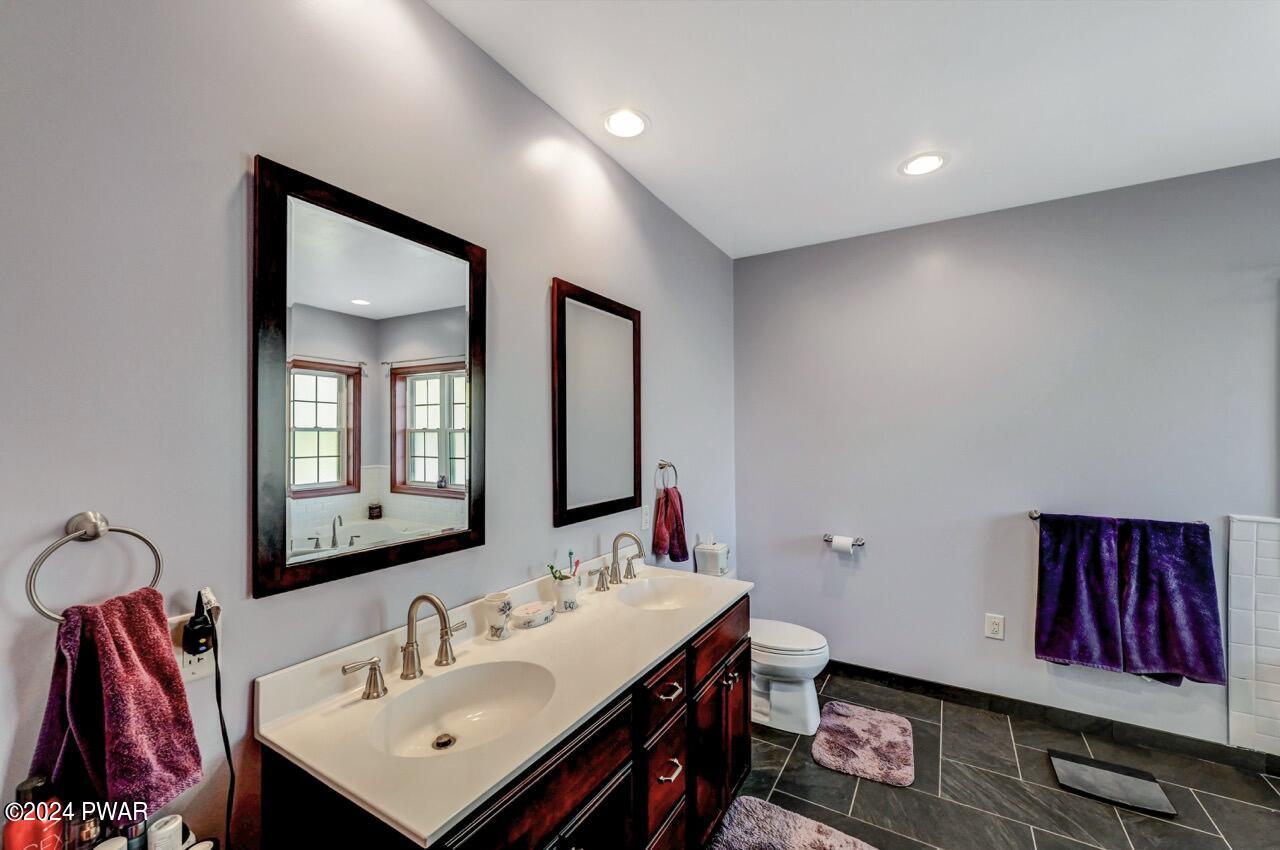 A modern bathroom with a double-sink vanity, large mirrors, recessed lighting, dark tile floor, and a toilet area with purple towels and bath mats.