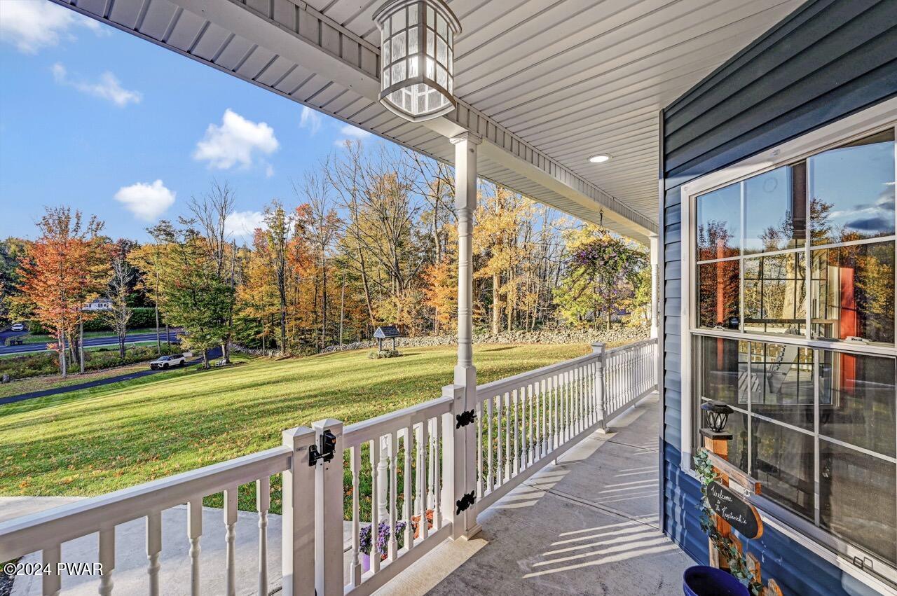 Covered front porch with white railing overlooking a grassy yard and autumn trees, with the house exterior and a large window on the right.