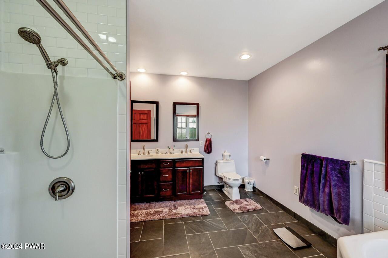 Spacious bathroom with a walk-in shower on the left, vanity with double sinks, toilet, dark tiled floor, and purple towels.
