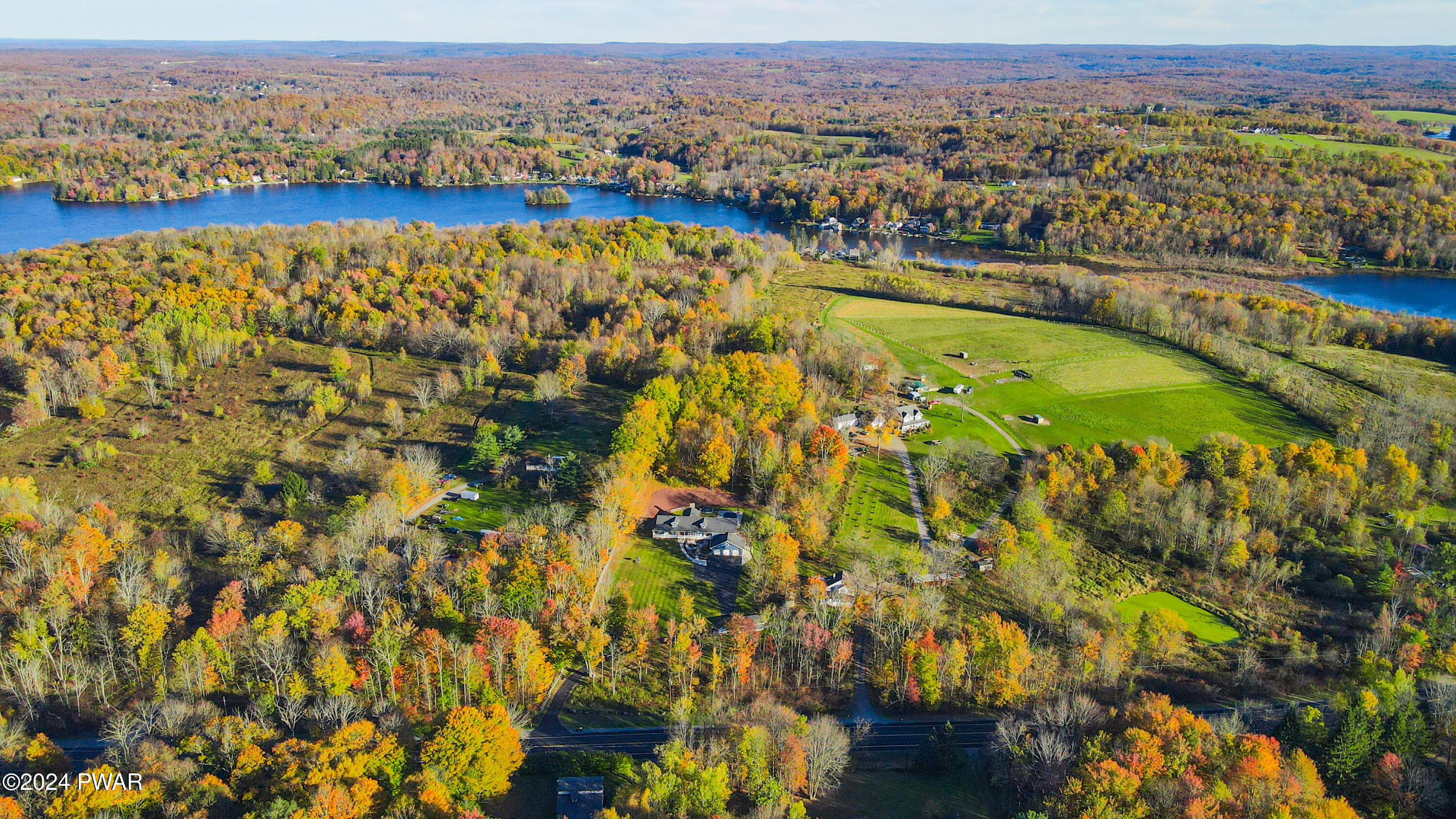 Aerial view of a rural lakeside landscape with autumn-colored trees, scattered homes, fields, and a large lake.