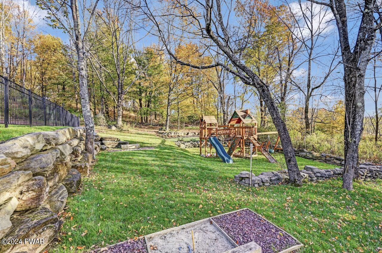 Landscaped backyard with a wooden playset and slide, stone retaining walls, trees, and green lawn in autumn.
