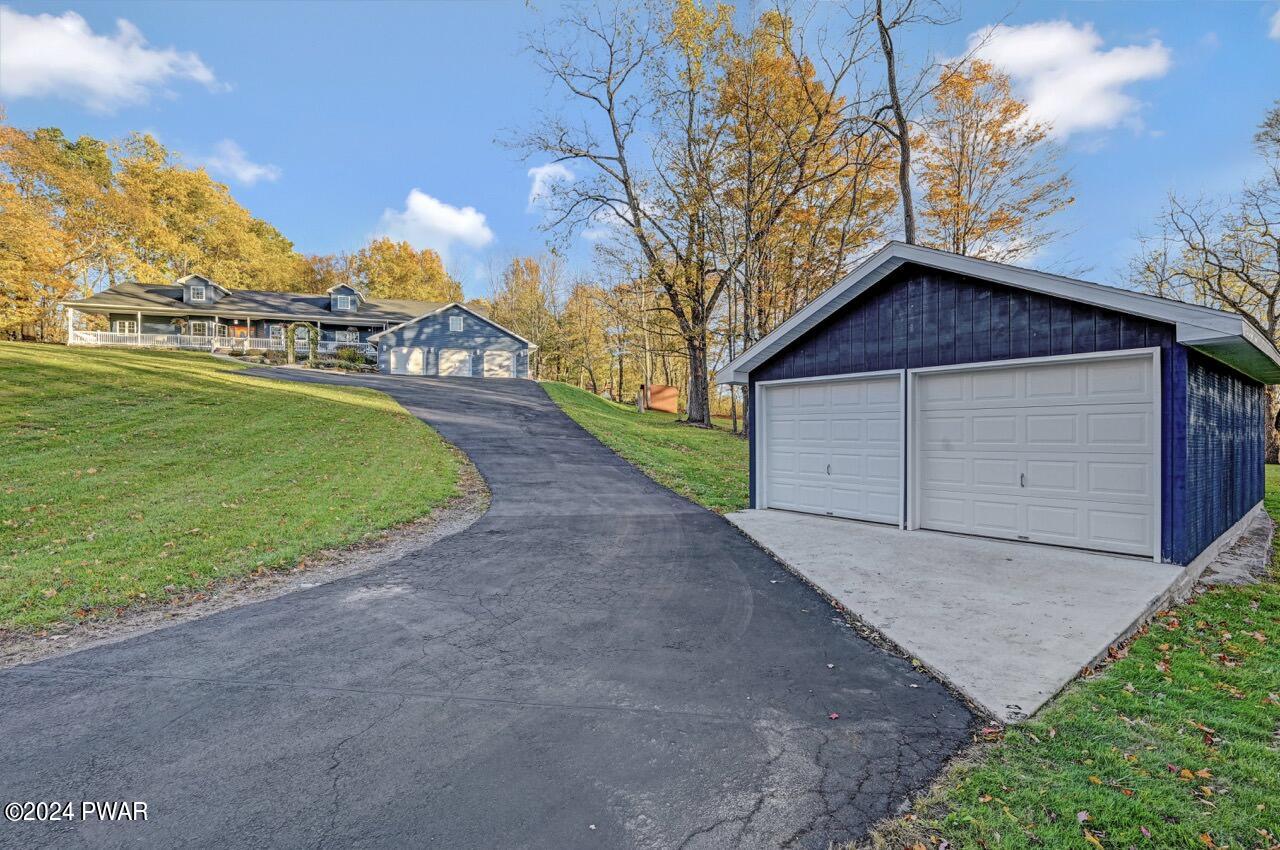 A paved driveway curves uphill toward a house on a grassy lot, with a detached two-car garage in the foreground and autumn trees in the background.