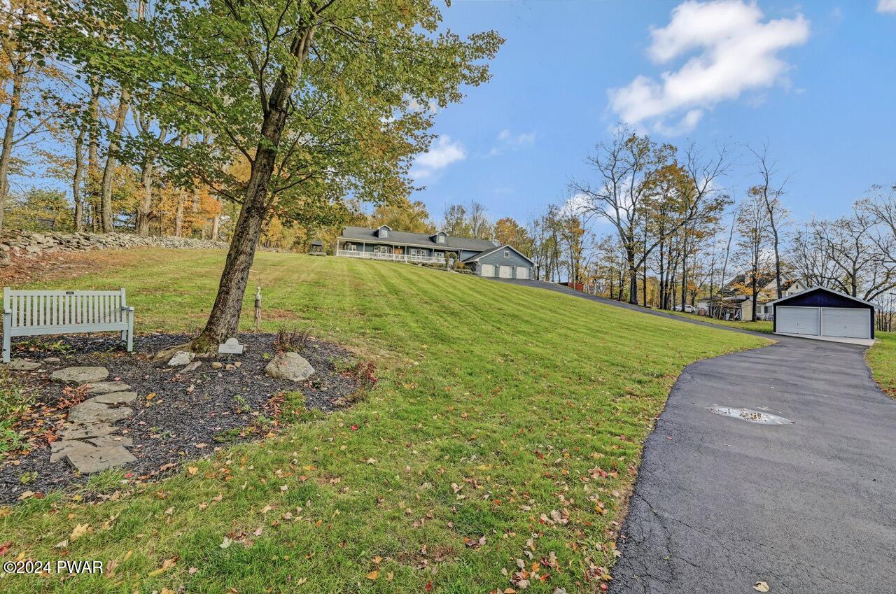 Wide exterior view of a sloped grassy yard with a driveway leading up to a house and detached garage under a blue sky.