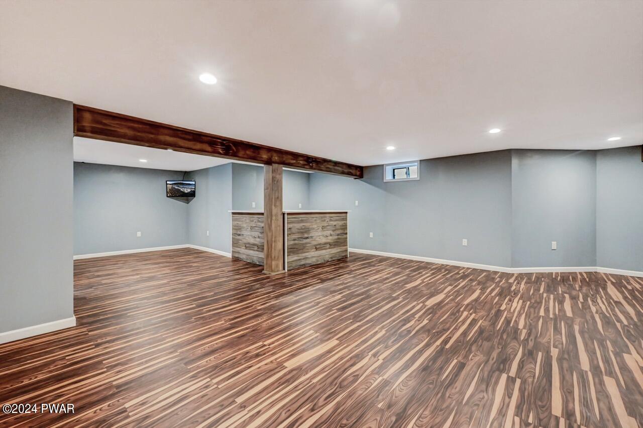 Empty finished basement with wood-look floors, gray walls, recessed lighting, exposed beam, and a small wet bar.