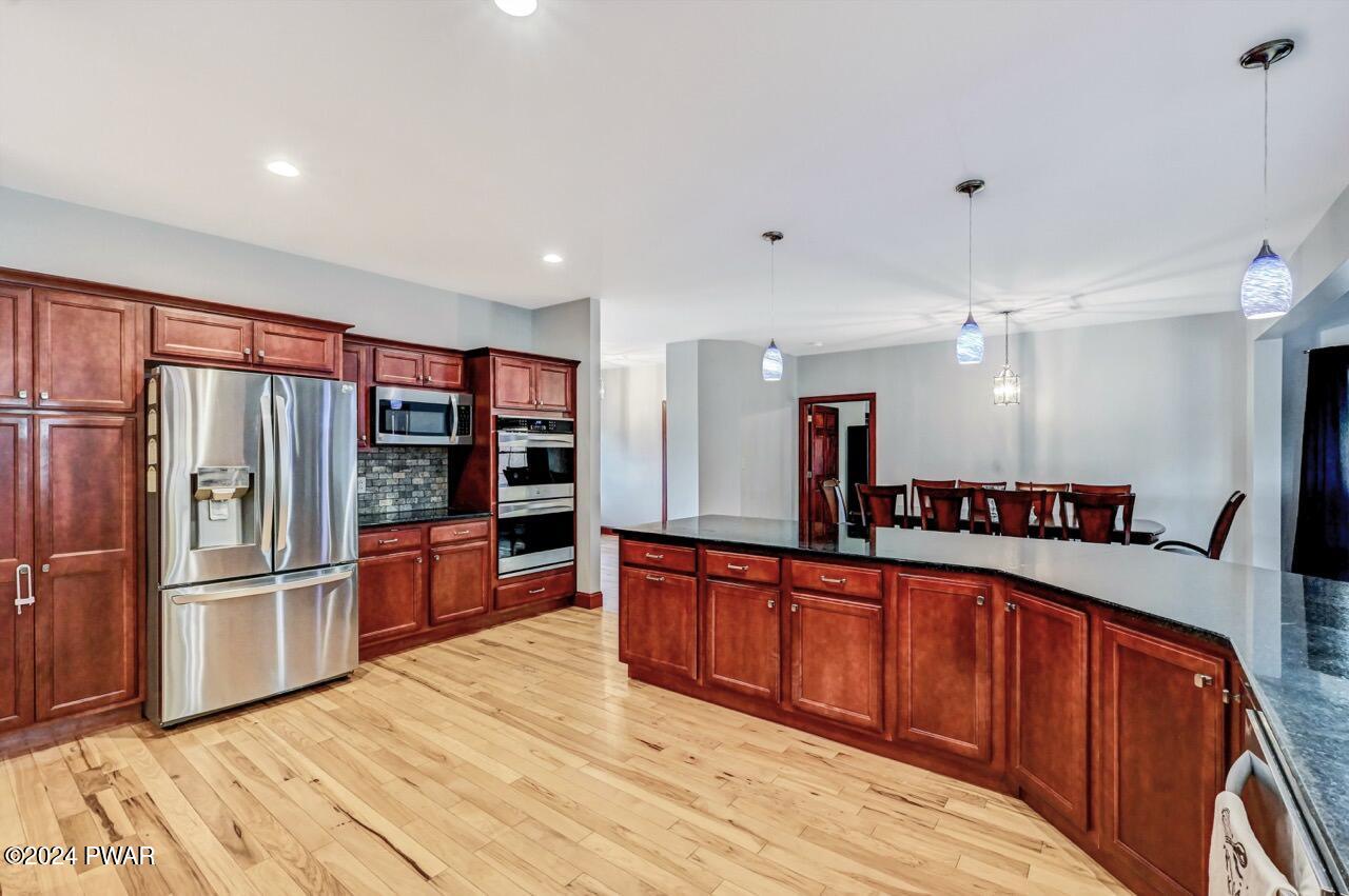 Spacious kitchen with hardwood floors, cherry cabinets, stainless steel refrigerator and built-in ovens, a large curved island, and a dining area in the background.